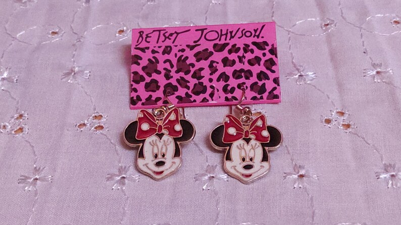 Pu&ograve; includere: Orecchini di Minnie Mouse con fiocco rosso e viso bianco, appesi a un cartoncino rosa con "Betsey Johnson" e stampa leopardata. Un accessorio divertente.