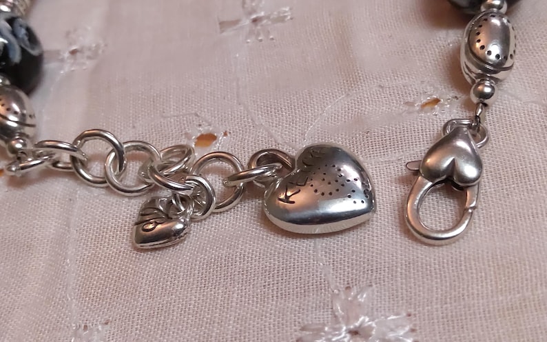 May include: Silver-toned bracelet with heart-shaped charms and a lobster clasp. The bracelet features a series of round links and decorative beads. The heart charms have a textured surface with small cut-out designs.
