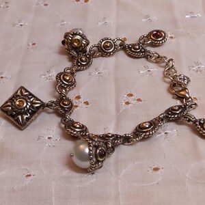 May include: Silver-toned charm bracelet with ornate detailing. Features a square charm, round charms with gold accents, and a dangling pearl. The bracelet has a lobster clasp closure and is set against a white, textured background.
