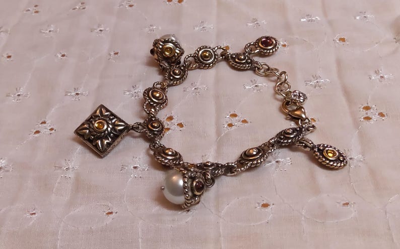 May include: Silver-toned charm bracelet with ornate detailing. Features a square charm, round charms with gold accents, and a dangling pearl. The bracelet has a lobster clasp closure and is set against a white, textured background.