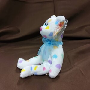 May include: A white plush beanie baby bear with colorful polka dots and a blue ribbon around its neck. The bear is sitting on a brown surface and has a Ty tag attached to its ear.