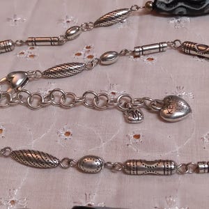 Op de afbeelding: Zilverkleurige ketting met diverse decoratieve kralen en bedels. De ketting heeft een hartvormige bedel met het woord "Keep" gegraveerd, kleine bedels en een karabijnsluiting. De kralen hebben gestructureerde patronen.