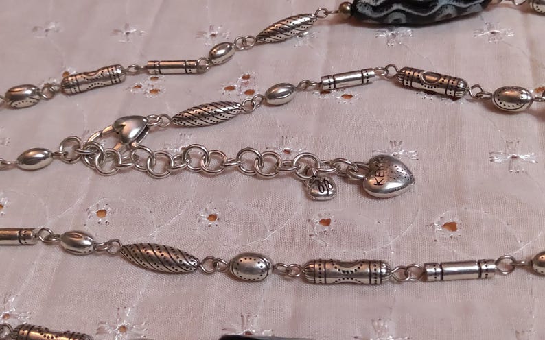 Op de afbeelding: Zilverkleurige ketting met diverse decoratieve kralen en bedels. De ketting heeft een hartvormige bedel met het woord "Keep" gegraveerd, kleine bedels en een karabijnsluiting. De kralen hebben gestructureerde patronen.