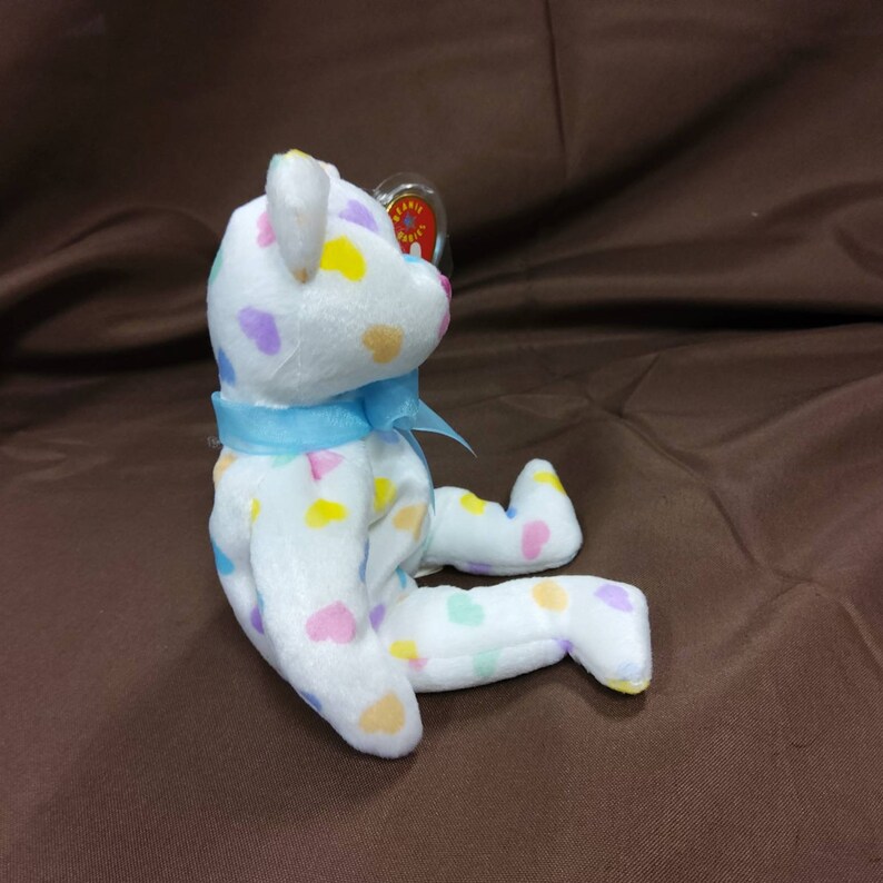 May include: White plush teddy bear with a blue ribbon around its neck. The bear is covered in colorful heart-shaped spots.