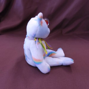 May include: A blue and white rainbow swirl beanie baby with a purple, yellow, and green ribbon around its neck. The beanie baby is sitting on a purple surface.