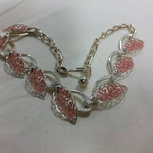 May include: A silver necklace with pink filigree detailing. The necklace is made up of a chain with several decorative links in the shape of leaves.