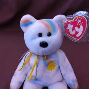 May include: A white rainbow beanie baby with a yellow sun on its chest and a purple, yellow, and green ribbon around its neck. The beanie baby has a Ty tag attached to its ear that says "Beanie Baby".