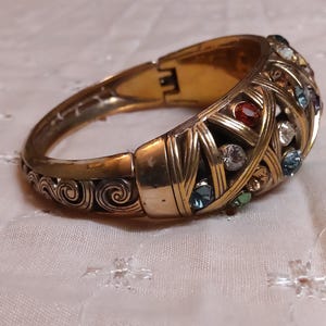 May include: A gold-toned hinged bracelet with a woven design, adorned with colorful gemstones. The bracelet features red, blue, green, and clear stones. The clasp is visible, and the bracelet rests on a white, patterned surface.