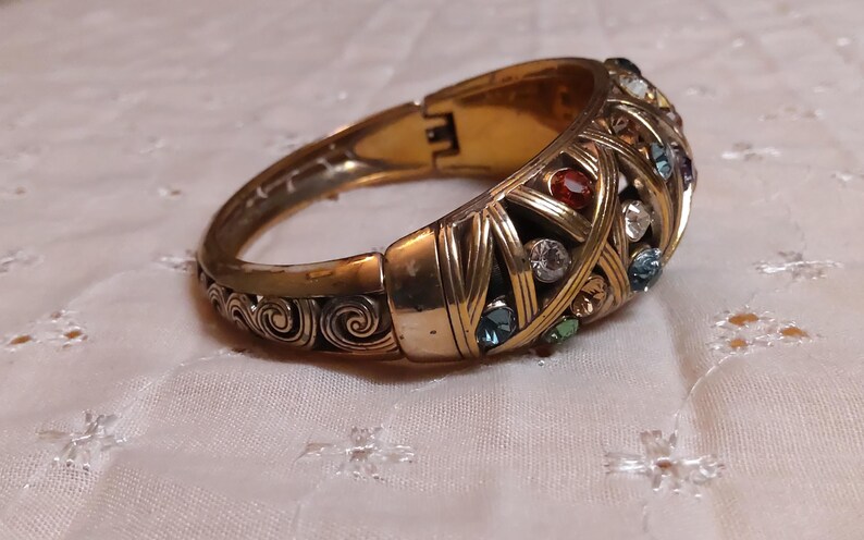 May include: A gold-toned hinged bracelet with a woven design, adorned with colorful gemstones. The bracelet features red, blue, green, and clear stones. The clasp is visible, and the bracelet rests on a white, patterned surface.