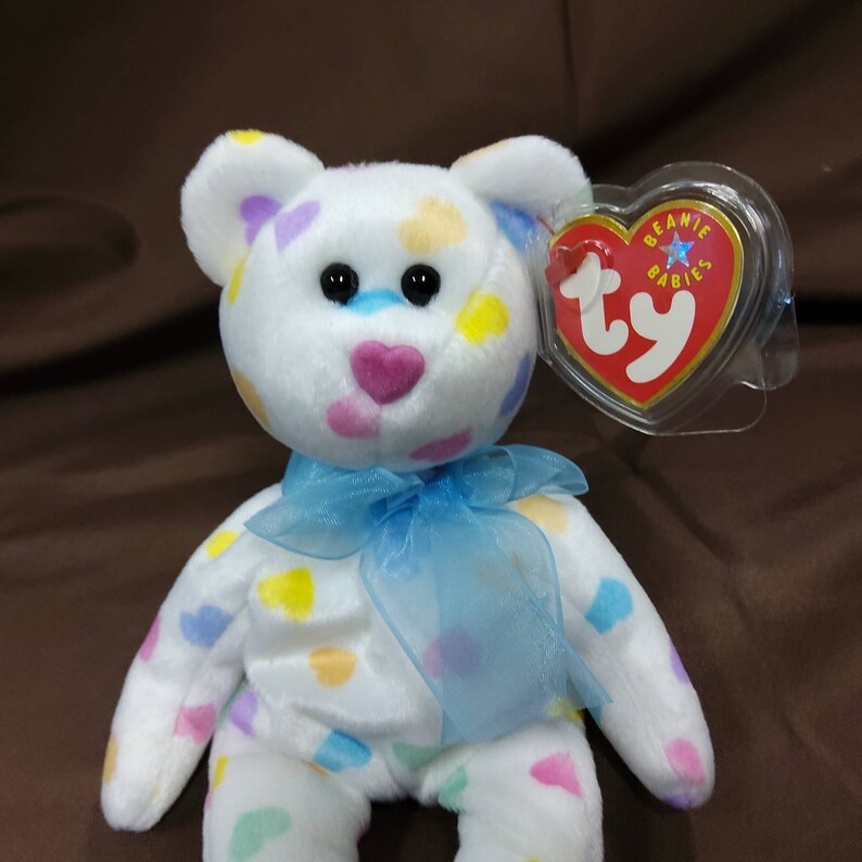 May include: White plush beanie baby bear with a blue ribbon around its neck. The bear is covered in colorful heart-shaped spots. The bear has a pink heart-shaped nose and black eyes. The bear has a Ty Beanie Babies tag attached to its ear.