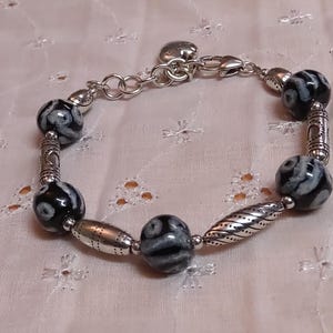 May include: A silver-toned bracelet featuring black and white patterned beads and decorative metal accents. The bracelet has a chain closure for adjustable sizing. The beads are approximately 0.75 inches in diameter.