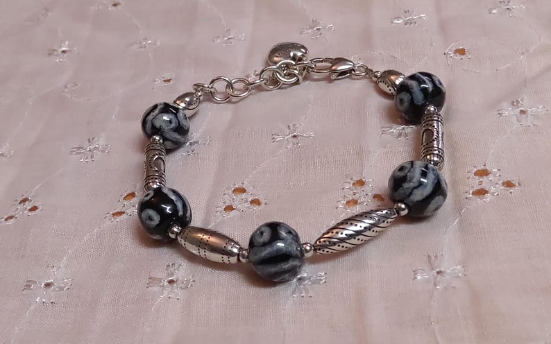 May include: A silver-toned bracelet featuring black and white patterned beads and decorative metal accents. The bracelet has a chain closure for adjustable sizing. The beads are approximately 0.75 inches in diameter.