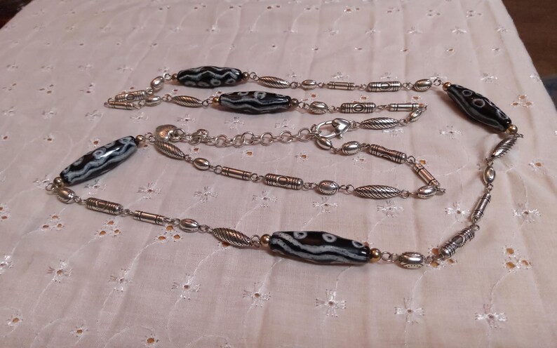 Op de afbeelding: Een lange ketting met afwisselend zwart-wit patronen kralen en zilverkleurige metalen schakels. De ketting heeft ovale, cilindrische en bladvormige metalen elementen, met een karabijnsluiting. De ketting wordt gepresenteerd op een wit, getextureerd oppervlak.