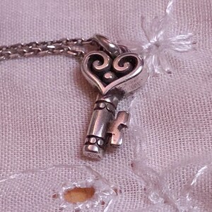 Op de afbeelding: Een zilverkleurige sleutelhangerketting met een hartvormige bovenkant en een sleutelgatontwerp. De sleutel hangt aan een fijne ketting. De ketting wordt gepresenteerd op een getextureerde, lichtroze stof.