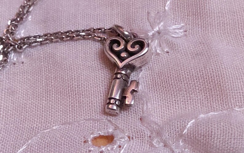 Op de afbeelding: Een zilverkleurige sleutelhangerketting met een hartvormige bovenkant en een sleutelgatontwerp. De sleutel hangt aan een fijne ketting. De ketting wordt gepresenteerd op een getextureerde, lichtroze stof.