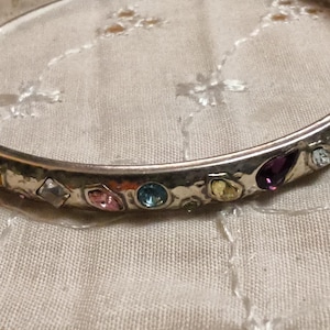 May include: A silver-toned bangle bracelet with a hammered texture. The bracelet is adorned with a variety of colorful gemstones, including purple, pink, blue, and yellow. The gemstones are set in individual bezels around the bracelet.
