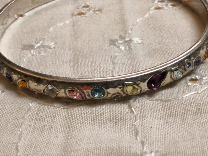 May include: A silver-toned bangle bracelet with a hammered texture. The bracelet is adorned with a variety of colorful gemstones, including purple, pink, blue, and yellow. The gemstones are set in individual bezels around the bracelet.