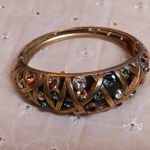 May include: Gold-toned hinged bangle bracelet with a woven design. The bracelet is adorned with a variety of colorful, sparkling gemstones, including clear, blue, green, and red stones. The bracelet is approximately 2.5 inches in diameter.