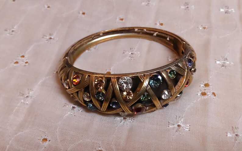 May include: Gold-toned hinged bangle bracelet with a woven design. The bracelet is adorned with a variety of colorful, sparkling gemstones, including clear, blue, green, and red stones. The bracelet is approximately 2.5 inches in diameter.