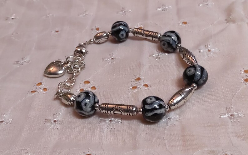 May include: A silver bracelet featuring black and white patterned beads and decorative silver accents. The bracelet includes a heart-shaped charm and is displayed on a textured, light pink surface.
