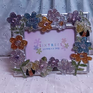 May include: A rectangular silver picture frame decorated with colorful glittery flowers. The frame holds a pink insert with the text "SIX TREES" and the dimensions 3.5 inch x 5 inch.