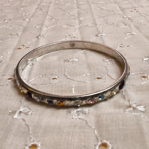 May include: Silver-toned bangle bracelet with colorful gemstones. The bracelet features a row of small, multi-colored stones set along the outer edge. A small heart-shaped design is visible on the bracelet. The bracelet is resting on a white, patterned surface.
