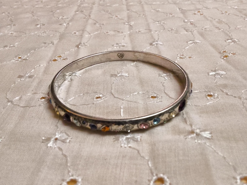 May include: Silver-toned bangle bracelet with colorful gemstones. The bracelet features a row of small, multi-colored stones set along the outer edge. A small heart-shaped design is visible on the bracelet. The bracelet is resting on a white, patterned surface.