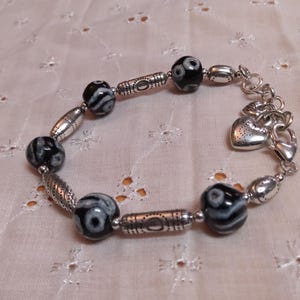 May include: A beaded bracelet featuring black and white patterned beads and silver-toned metal accents. The bracelet includes decorative oval and cylindrical beads, and a heart-shaped charm. The bracelet is displayed on a light pink, patterned fabric.