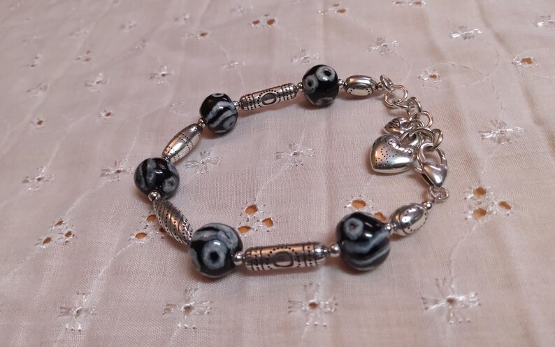 May include: A beaded bracelet featuring black and white patterned beads and silver-toned metal accents. The bracelet includes decorative oval and cylindrical beads, and a heart-shaped charm. The bracelet is displayed on a light pink, patterned fabric.