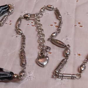 Op de afbeelding: Een zilveren ketting met een verscheidenheid aan kralen en bedels. De ketting heeft grote, zwart-wit patronen kralen, kleinere zilveren kralen en decoratieve zilveren buizen. Een hartvormige bedel hangt in de buurt van de sluiting.