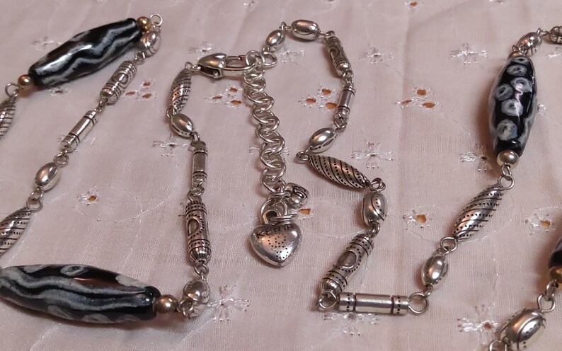 Op de afbeelding: Een zilveren ketting met een verscheidenheid aan kralen en bedels. De ketting heeft grote, zwart-wit patronen kralen, kleinere zilveren kralen en decoratieve zilveren buizen. Een hartvormige bedel hangt in de buurt van de sluiting.
