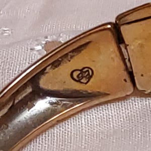 May include: Close-up of a gold-toned bracelet segment. The bracelet has a heart-shaped emblem with an intertwined design. The metal has a slightly aged appearance. The bracelet is on a white, textured surface.
