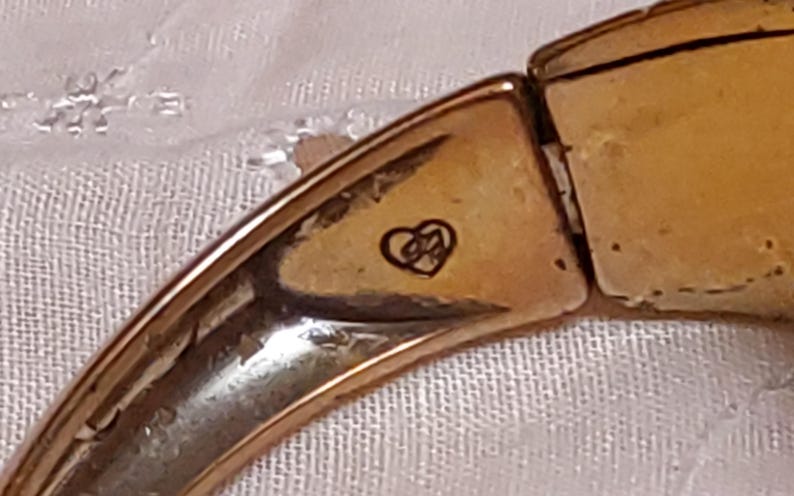 May include: Close-up of a gold-toned bracelet segment. The bracelet has a heart-shaped emblem with an intertwined design. The metal has a slightly aged appearance. The bracelet is on a white, textured surface.