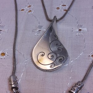 May include: A silver necklace with a teardrop-shaped pendant featuring a swirling design. The pendant is attached to a silver chain.