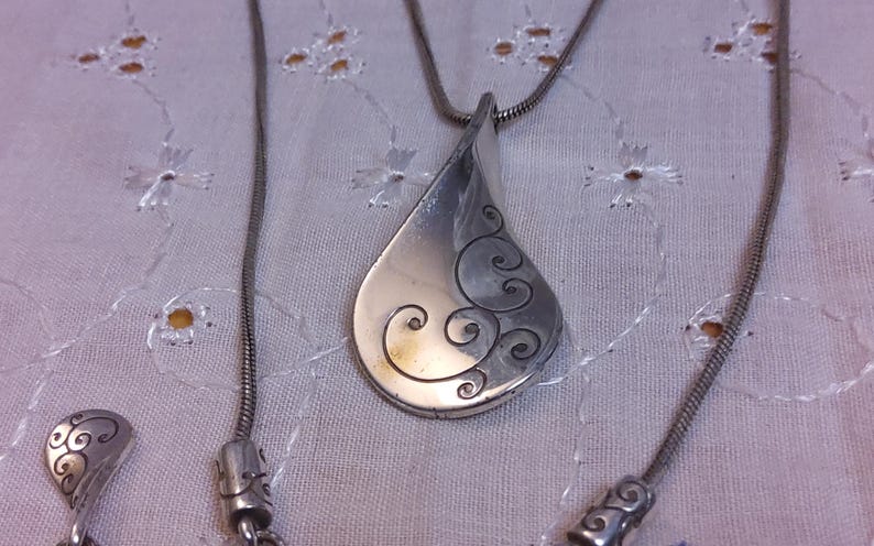 May include: A silver necklace with a teardrop-shaped pendant featuring a swirling design. The pendant is attached to a silver chain.