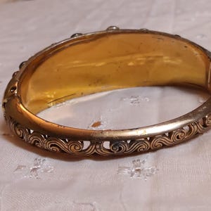 May include: A gold-toned hinged bangle bracelet. The interior is a bright gold color, while the exterior features an ornate, openwork design along the edges. The bracelet is resting on a white, patterned surface.
