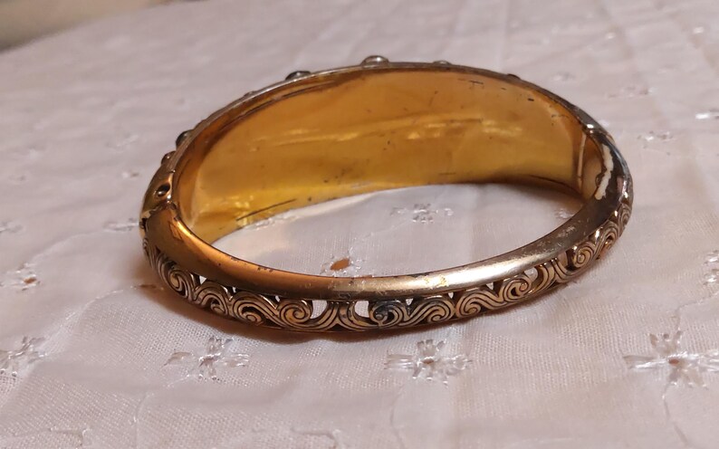 May include: A gold-toned hinged bangle bracelet. The interior is a bright gold color, while the exterior features an ornate, openwork design along the edges. The bracelet is resting on a white, patterned surface.