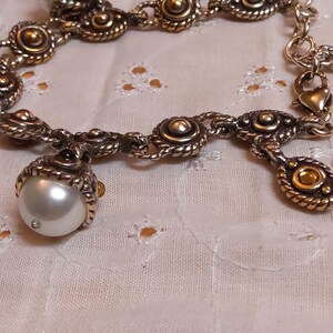 May include: A silver-toned bracelet with ornate detailing. The bracelet features a large, pearlescent white pearl charm and several smaller decorative elements with gold accents. The design includes rope-like textures and circular embellishments.