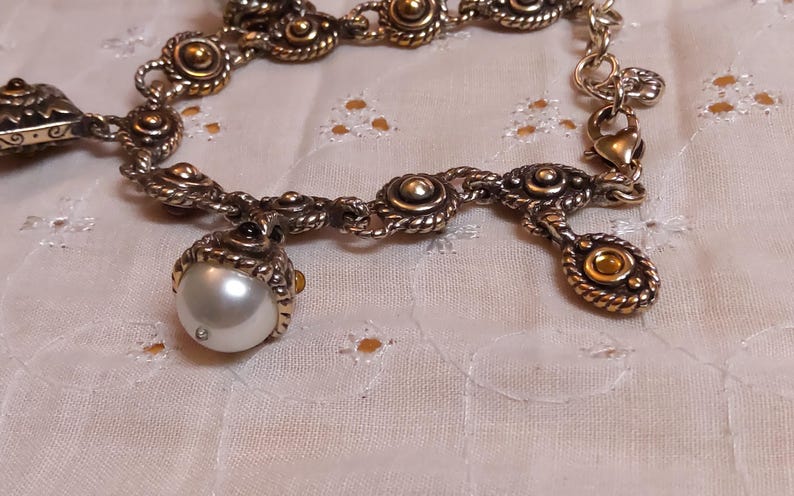 May include: A silver-toned bracelet with ornate detailing. The bracelet features a large, pearlescent white pearl charm and several smaller decorative elements with gold accents. The design includes rope-like textures and circular embellishments.