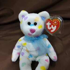 May include: White plush beanie baby bear with a blue ribbon around its neck. The bear is covered in colorful heart-shaped patches. The bear has a Ty tag attached to its ear that reads "Beanie Babies".