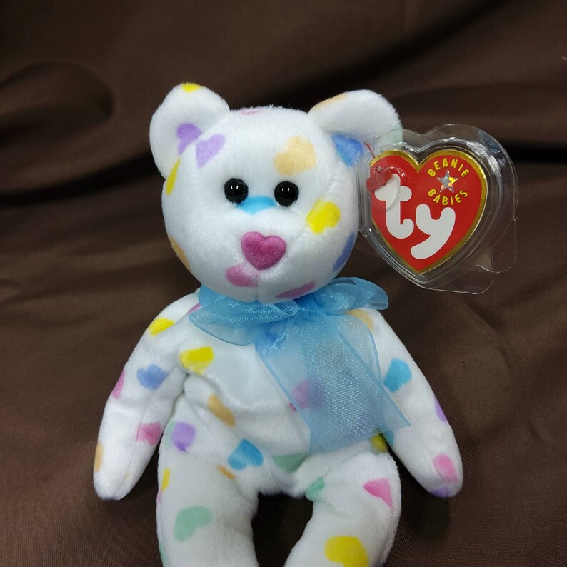 May include: White plush beanie baby bear with a blue ribbon around its neck. The bear is covered in colorful heart-shaped patches. The bear has a Ty tag attached to its ear that reads "Beanie Babies".