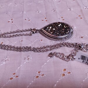 Może przedstawiać: Srebrny naszyjnik z wisiorkiem w kształcie łzy wypełnionym ciemnymi, błyszczącymi kamieniami i ozdobną srebrną obw&oacute;dką. Łańcuszek ma podw&oacute;jną konstrukcję z zapięciem na karabińczyk. Dołączona jest metka z ceną.