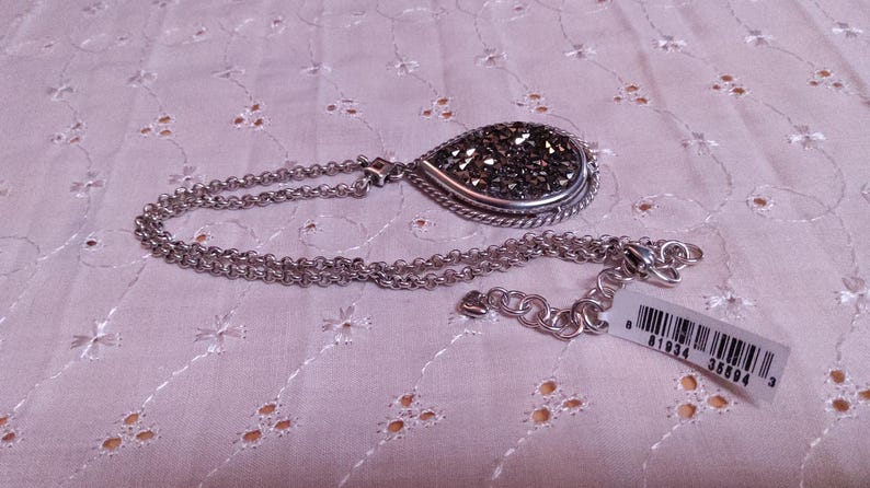 Może przedstawiać: Srebrny naszyjnik z wisiorkiem w kształcie łzy wypełnionym ciemnymi, błyszczącymi kamieniami i ozdobną srebrną obw&oacute;dką. Łańcuszek ma podw&oacute;jną konstrukcję z zapięciem na karabińczyk. Dołączona jest metka z ceną.