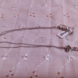 Op de afbeelding: Zilverkleurige ketting met een sleutelhanger. De sleutelhanger heeft een hartvormig ontwerp. De ketting ligt op een witte, geborduurde stof. Een label met het woord "Brighton" is bevestigd.