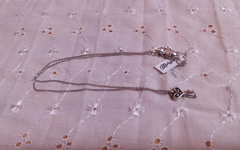 Op de afbeelding: Zilverkleurige ketting met een sleutelhanger. De sleutelhanger heeft een hartvormig ontwerp. De ketting ligt op een witte, geborduurde stof. Een label met het woord "Brighton" is bevestigd.