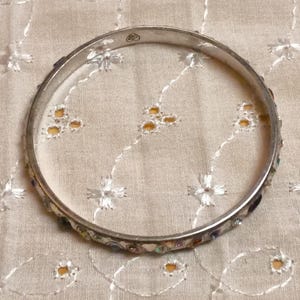 May include: A silver-toned bangle bracelet with colorful, small, inlaid stones. The bracelet is circular and rests on a cream-colored, embroidered fabric. The stones are various colors, including blue, red, and yellow.