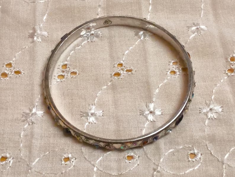 May include: A silver-toned bangle bracelet with colorful, small, inlaid stones. The bracelet is circular and rests on a cream-colored, embroidered fabric. The stones are various colors, including blue, red, and yellow.