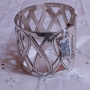 May include: Silver-toned metal cuff bracelet with an openwork design of intersecting ribbon shapes. The bracelet has a polished finish and a tag that reads "Brighton". The cuff is displayed on a white, textured surface.