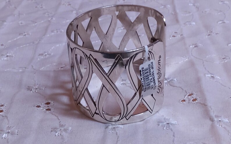 May include: Silver-toned metal cuff bracelet with an openwork design of intersecting ribbon shapes. The bracelet has a polished finish and a tag that reads "Brighton". The cuff is displayed on a white, textured surface.