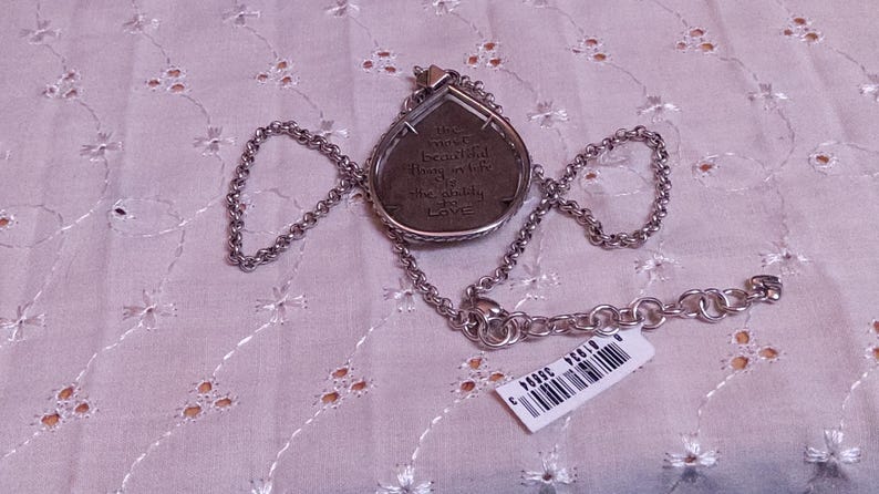 Może przedstawiać: Srebrny naszyjnik z wisiorkiem w kształcie łzy. Wisiorek ma napis i jest przymocowany do łańcuszka. Widoczna jest metka z ceną. Naszyjnik spoczywa na białej, wzorzystej powierzchni.